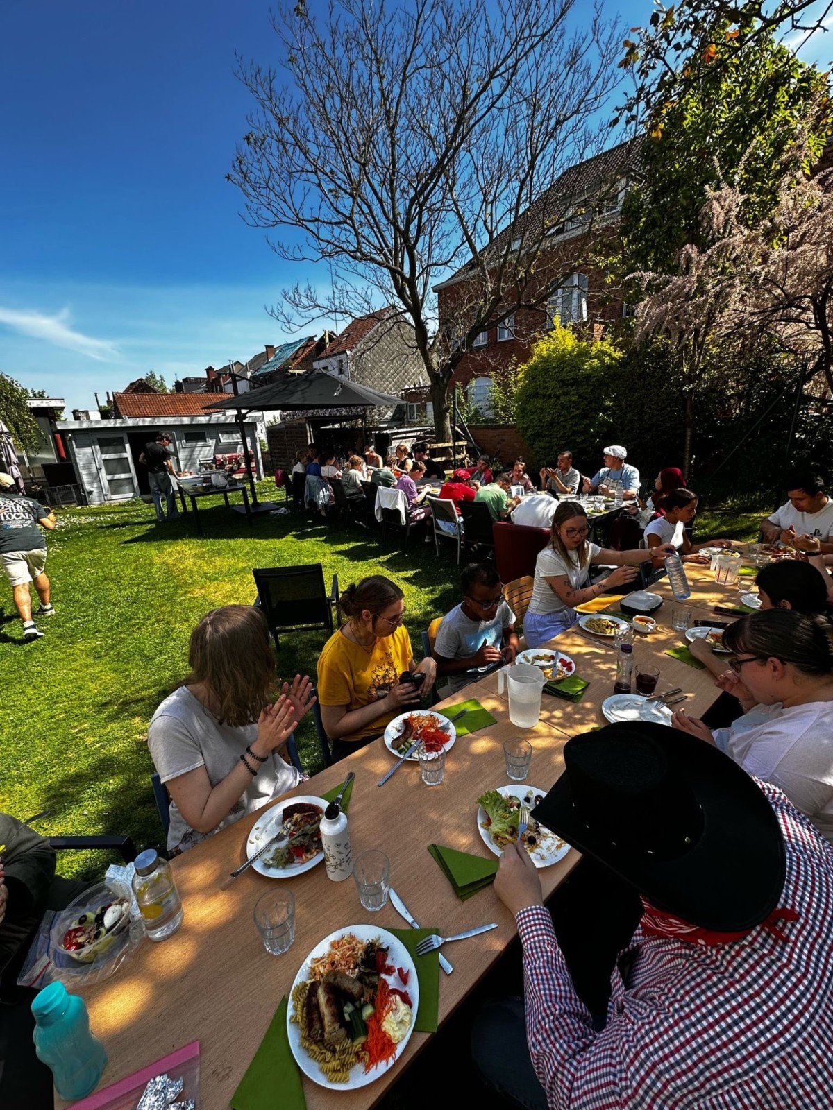Een groep mensen zit aan een lange tafel in een tuin, genietend van een maaltijd onder een heldere blauwe lucht, met verschillende borden vol eten voor zich.