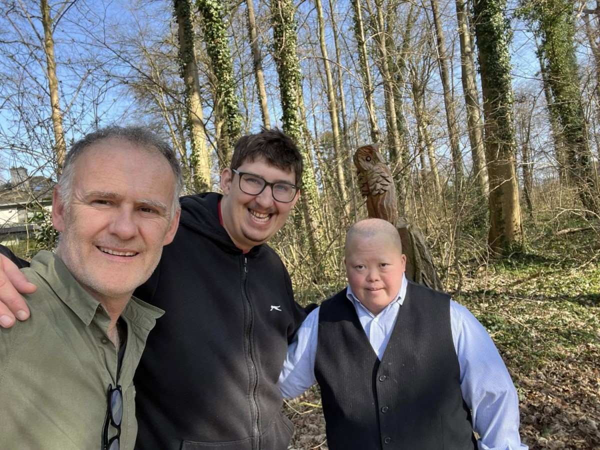 Drie mensen poseren samen voor een houtachtige achtergrond in een bos, met één persoon die een houten vogel in de achtergrond heeft.