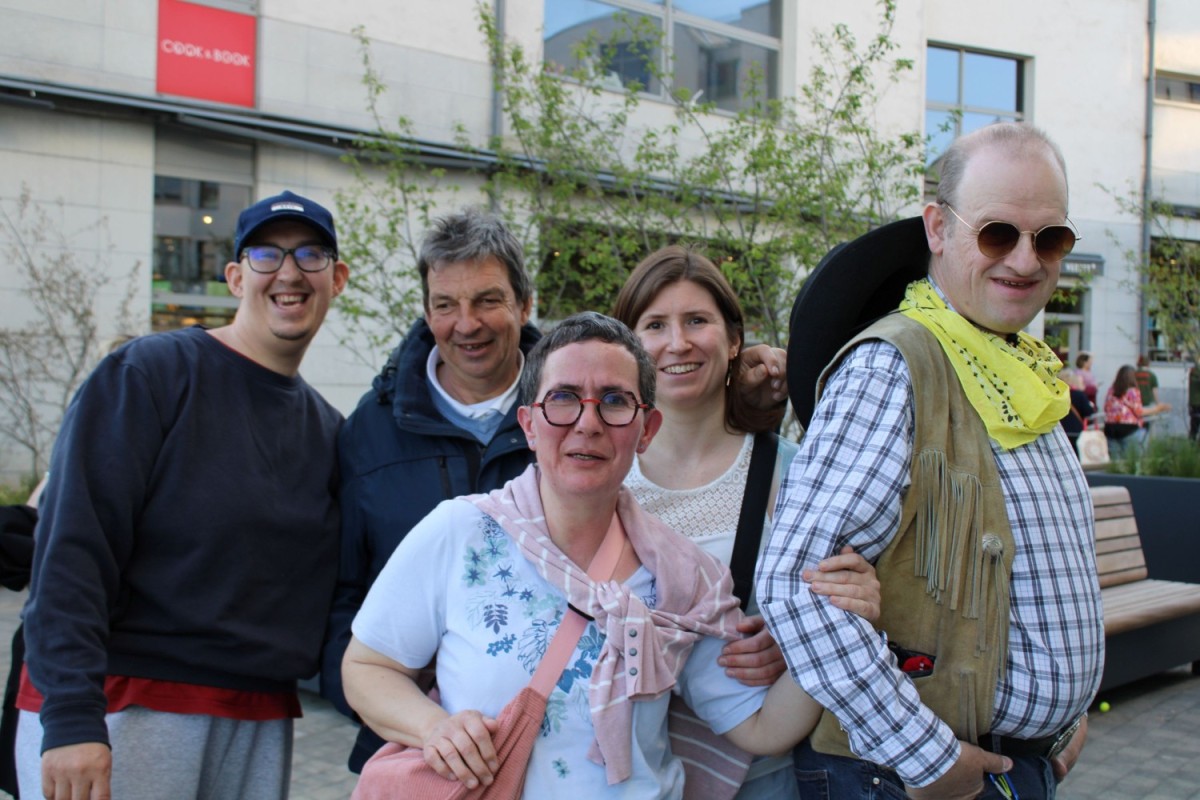 Een groep van vijf mensen poseert vrolijk voor een gebouw, met een groene begroeiing op de achtergrond.