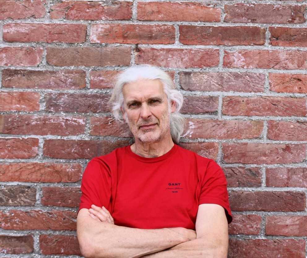Een man met lang, grijs haar in een rode T-shirt leunt met gekruiste armen tegen een bakstenen muur.