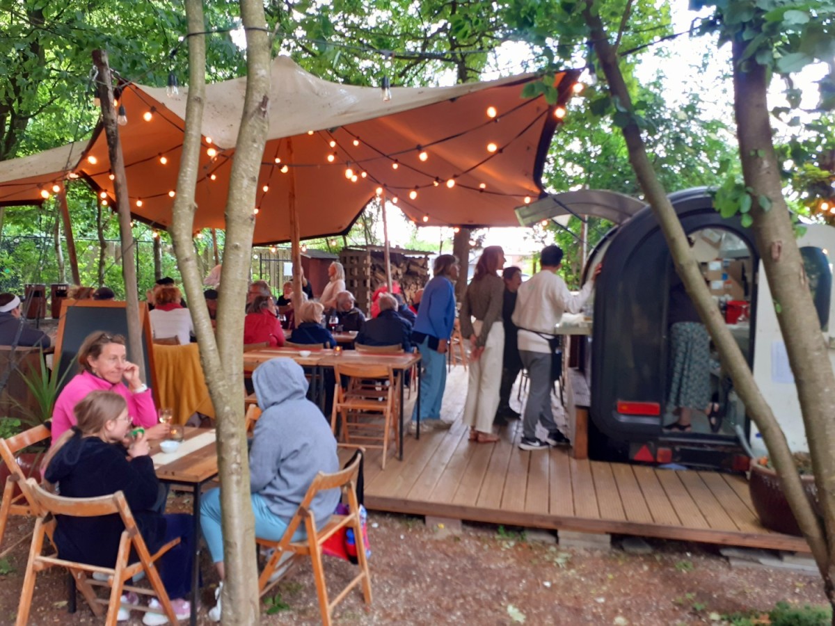 Gezellige buitenruimte met een grote tent, verwarmd door sfeervolle lampjes, waar gasten aan tafels zitten te genieten van hun maaltijden en anderen aan een eetkraam staan.