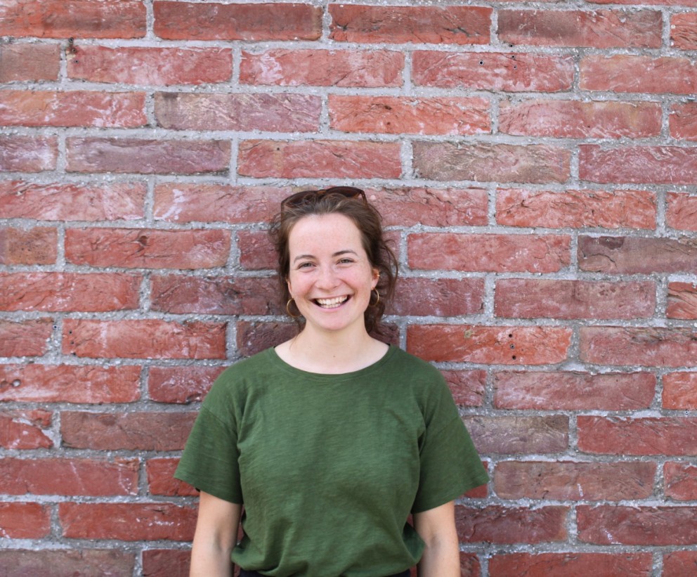 Een jonge vrouw met een glimlach staat tegen een bakstenen muur, gekleed in een groene T-shirt.