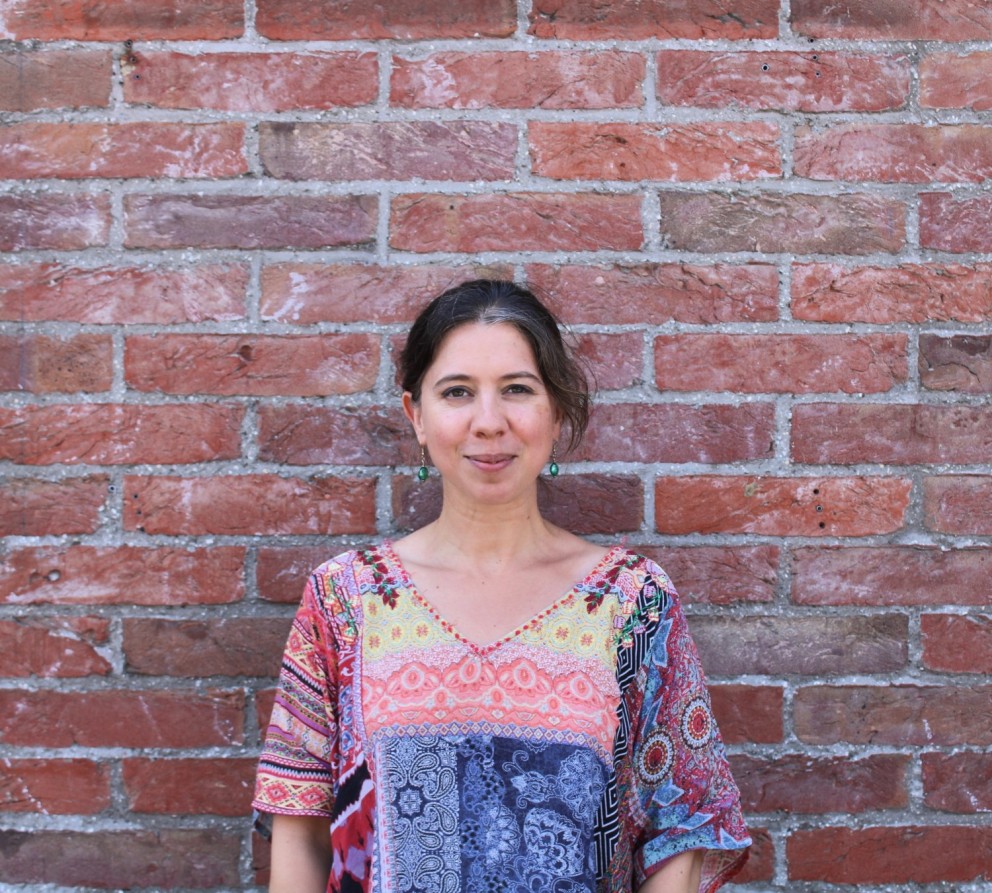 Vrouw in een kleurrijke blouse staat voor een bakstenen muur, met een rustige uitdrukking en loshangend haar.