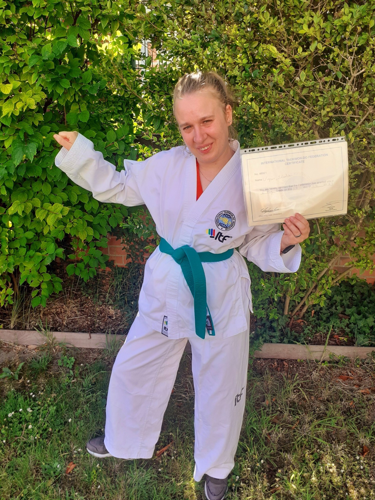 Een vrouw in een wit taekwondo-uniform met een groene band poseert trots in een tuin, terwijl ze een certificaat omhoog houdt.