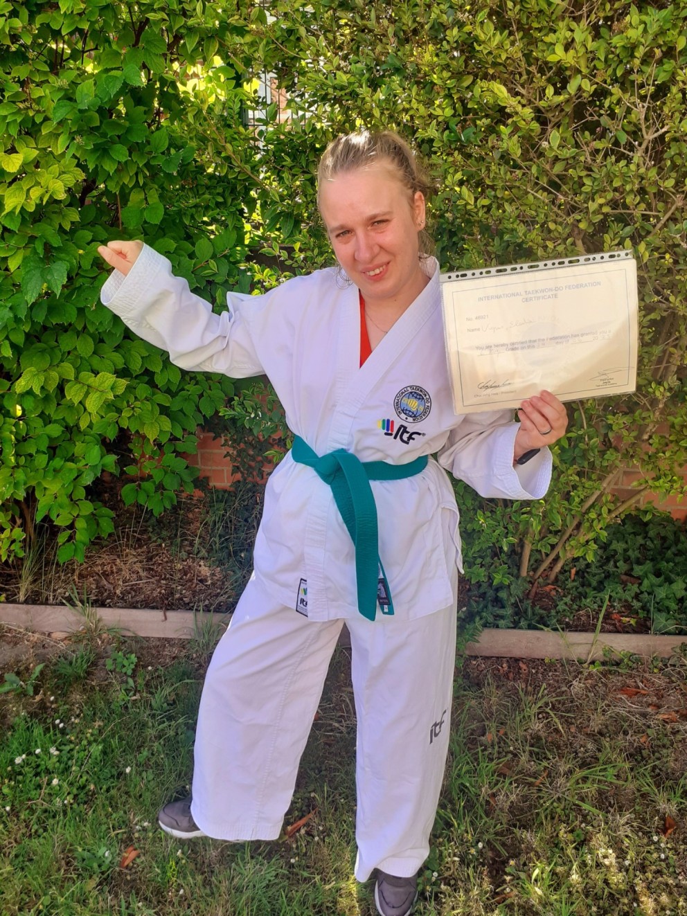 Een vrouw in een wit taekwondo-uniform met een groene band poseert trots in een tuin, terwijl ze een certificaat omhoog houdt.