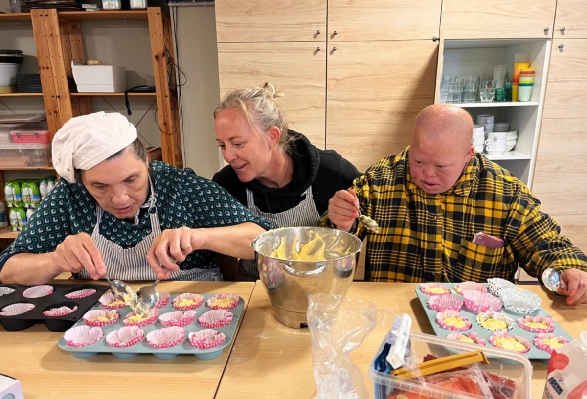 Drie mensen zijn aan het bakken in een keuken, waarbij ze beslag in cupcakevormpjes scheppen en genieten van het proces.