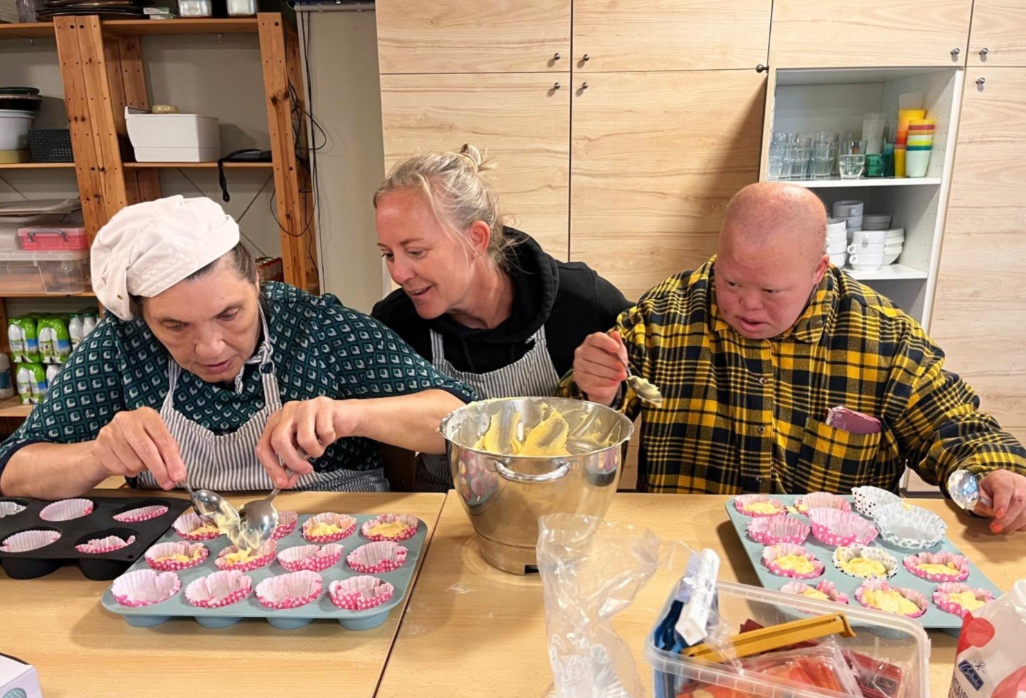 Drie mensen zijn aan het bakken in een keuken, waarbij ze beslag in cupcakevormpjes scheppen en genieten van het proces.