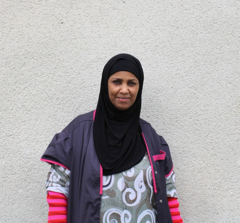 Een vrouw in een zwart hoofddoek en kleurrijke kleding staat tegen een lichte muur.