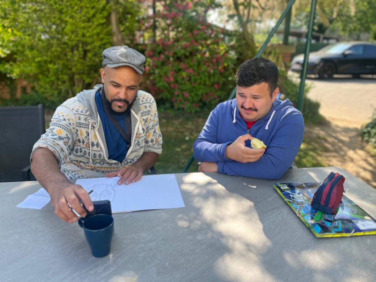 Twee mannen zitten aan een tafel in de buitenlucht; de man links, met een hoed, tekent en houdt een telefoon vast, terwijl de man rechts een stuk fruit vasthoudt en naar de andere man kijkt. Achter hen zijn bomen en bloemen zichtbaar.