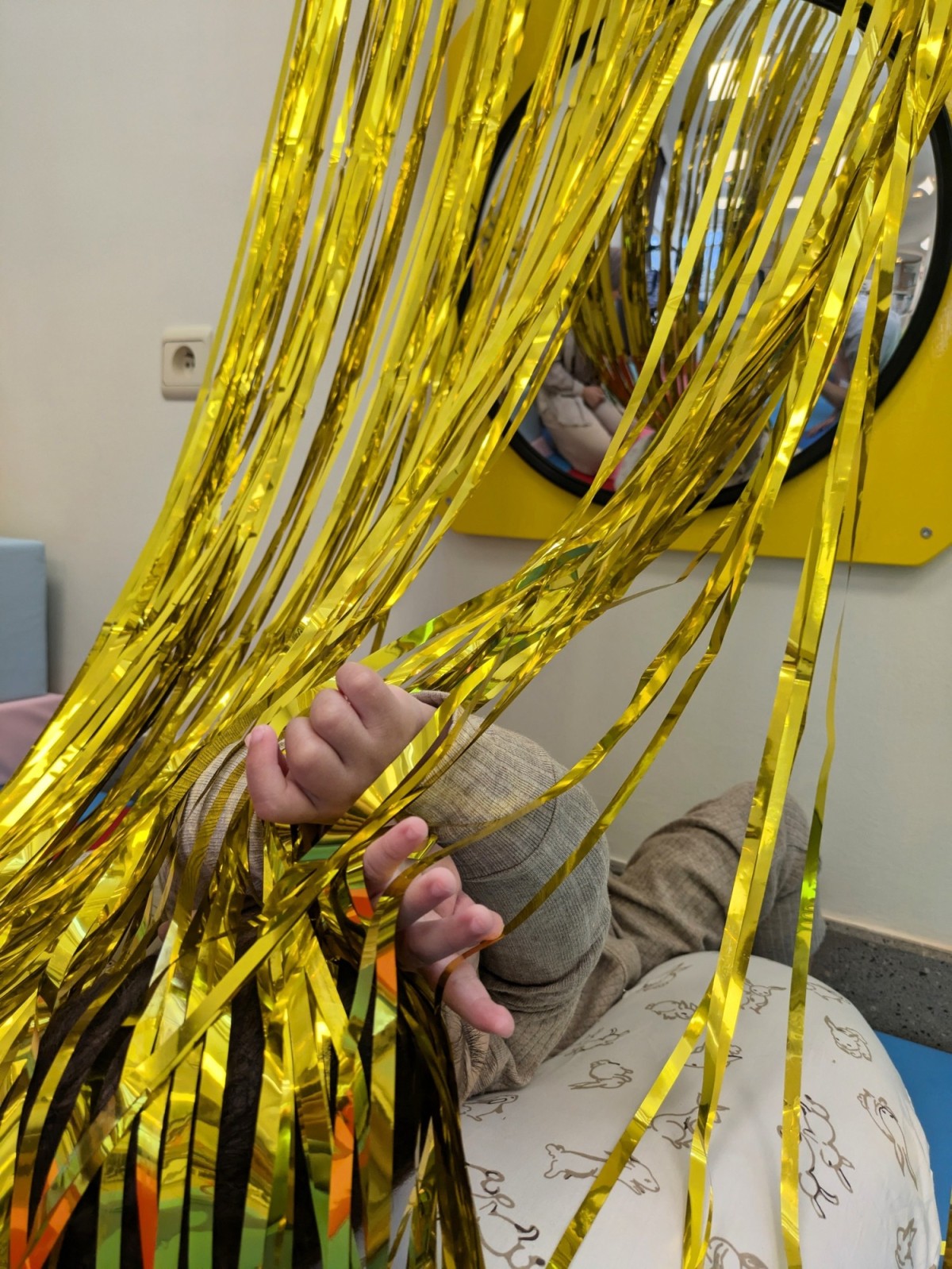 Een hand van een kind speelt met glanzende gouden strips in een speelhoek, met een ronde spiegel op de achtergrond.