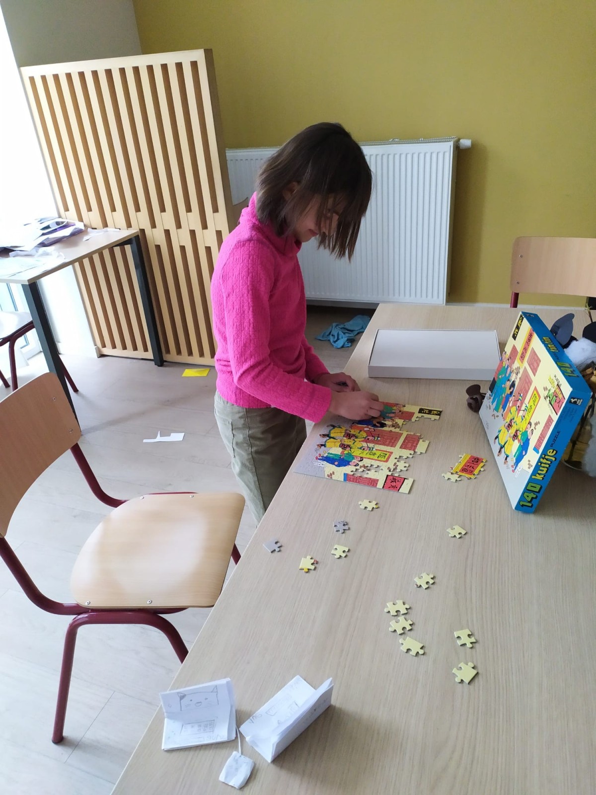 Een meisje in een roze trui dat een puzzel in elkaar zet op een houten tafel, met puzzelstukjes verspreid rondom haar. Achter haar staat een houten scherm en een verwarming.