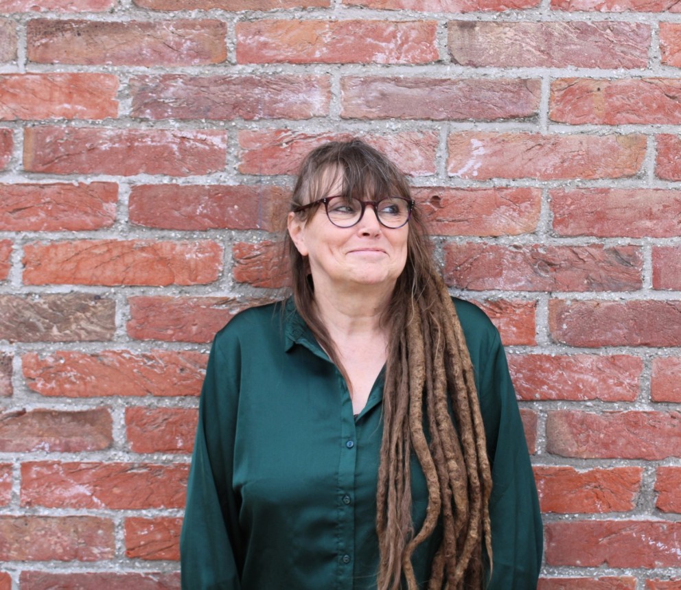 Vrouw met lang, dreadlock-haar in een groene blouse, staat met een glimlach tegen een bakstenen muur.