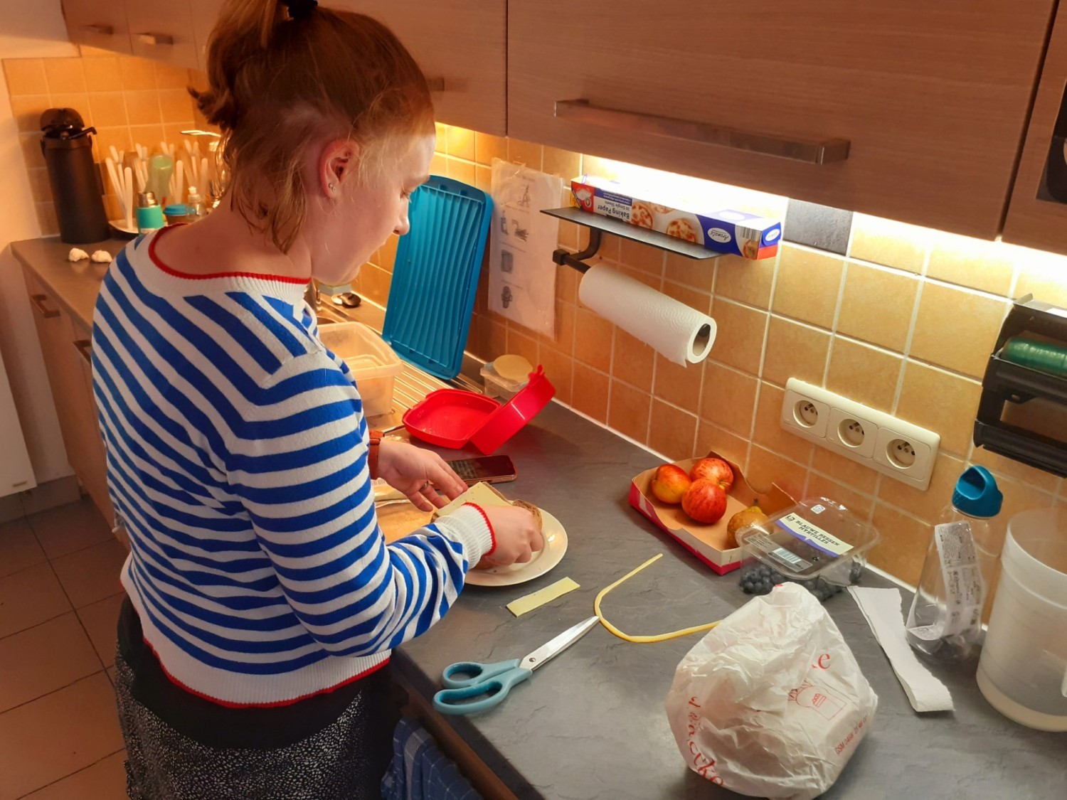 Een persoon met een gestreepte trui staat aan een keukenblad, waar ze aan het koken of voorbereiden is, omringd door verschillende gebruiksvoorwerpen en ingrediënten.