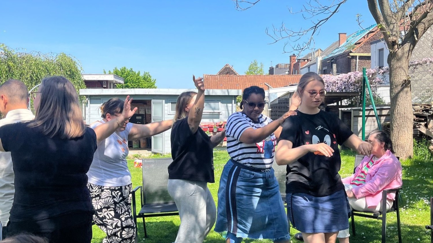 Vijf mensen dansen buiten in een groene omgeving, met een blauwe lucht en enkele gebouwen op de achtergrond.